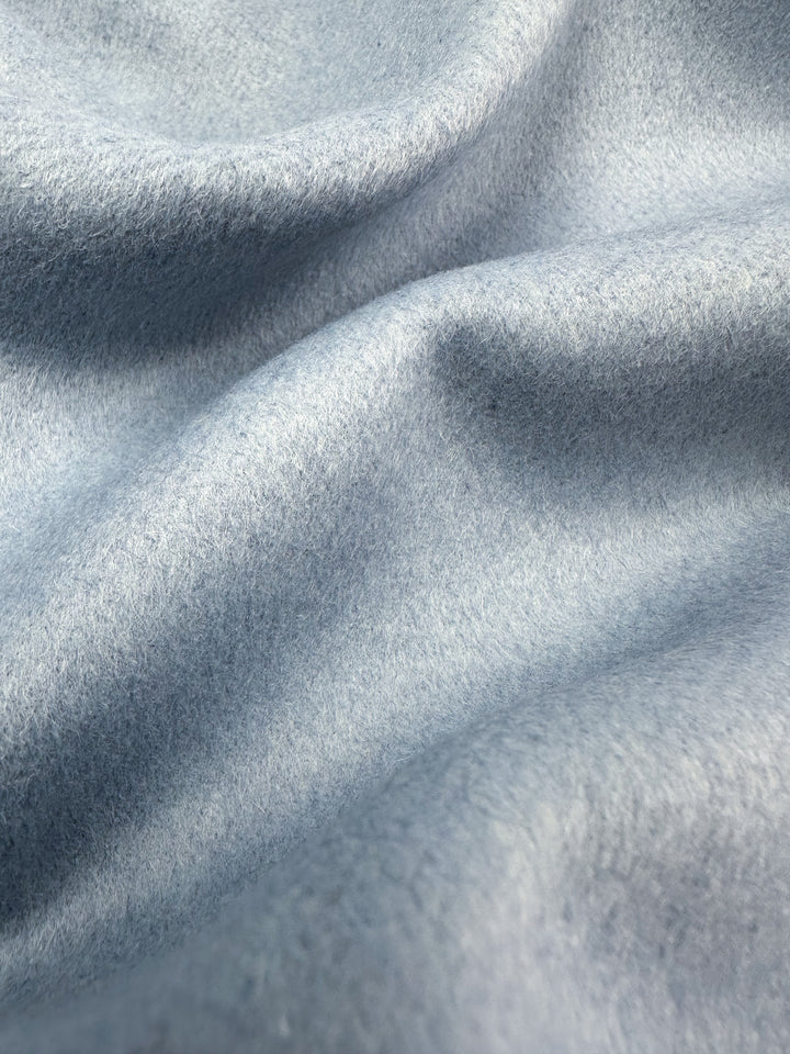 Close-up of Super Cheap Fabrics' Wool Cashmere - Niagara Mist (150cm): a soft, light blue fabric with gentle folds and a mildly fuzzy texture. Its elegant surface appears smooth, with subtle shadows enhancing the luxurious undulating pattern.