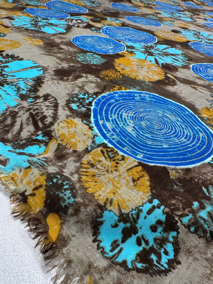 A close-up of the Super Cheap Fabrics' "Linen Blend - Radar On Earth - 145cm" showcases a lightweight texture with blue concentric circles, abstract floral patterns in yellow and turquoise, set against a brown and beige backdrop with fringed edges.