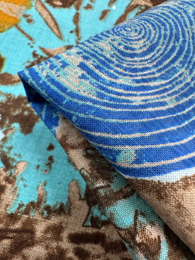 Close-up of the Linen Blend - Radar On Earth by Super Cheap Fabrics. This 145cm lightweight linen-cotton fabric displays a breathable, abstract design with blue and white spirals, plus brown and teal splatters. The textile is folded to highlight its vibrant, intricate pattern.