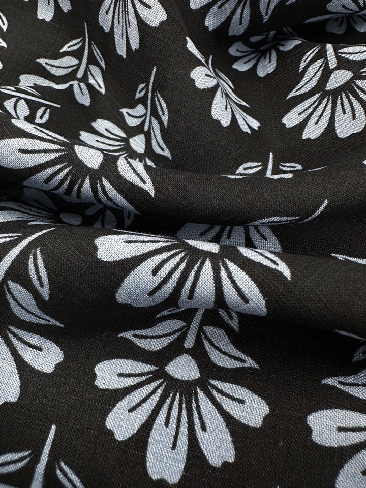 The Linen Blend - Chambray Floretta from Super Cheap Fabrics features a lightweight linen-cotton mix with a black base, adorned by evenly distributed abstract floral patterns. The breathable and soft fabric displays visible folds and textures across its 138cm width.