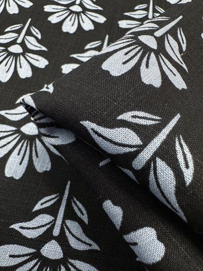 A close-up of the Linen Blend - Chambray Floretta - 138cm from Super Cheap Fabrics shows black breathable fabric with a repeated white floral pattern. This stylized design, crafted from lightweight linen cotton blend, features symmetrical flowers and is slightly folded at the center.