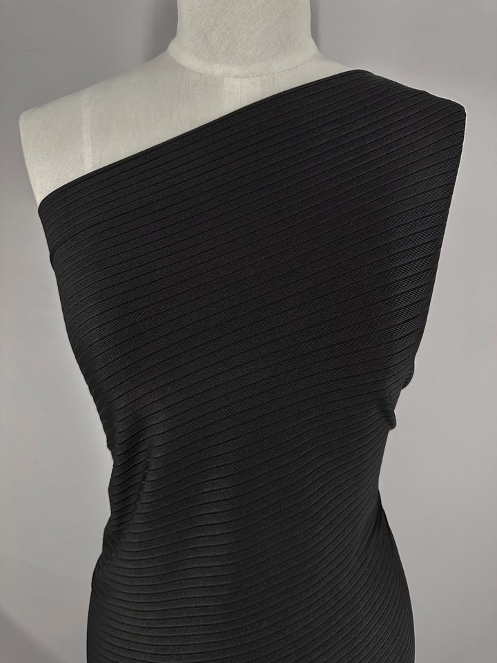 A mannequin showcases a black, one-shoulder dress crafted from Super Cheap Fabrics’ Rib Knit - Black (170cm), featuring medium weight and diagonal rib lines for a textured look. The stretchy material ensures comfort against a neutral gray backdrop.