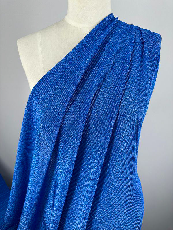 A close-up of a mannequin draped in Super Cheap Fabrics' Metallic Knit - Cobalt - 150cm. The medium to heavy weight material is pleated and flows elegantly over the mannequin's shoulder, showcasing its intricate, ribbed pattern. The background is a plain, light gray color.