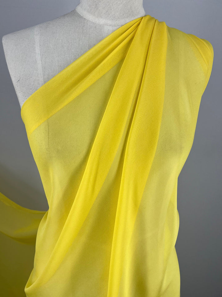 A mannequin draped in flowing, lightweight Silk Georgette - Quail from Super Cheap Fabrics is arranged in a stylish, asymmetrical manner. The sheer yellow material cascades gracefully over the shoulder, adding an elegant and ethereal look to the setup. Perfect for summer outfits, this 100% silk piece stands out against the plain background at 135cm wide.