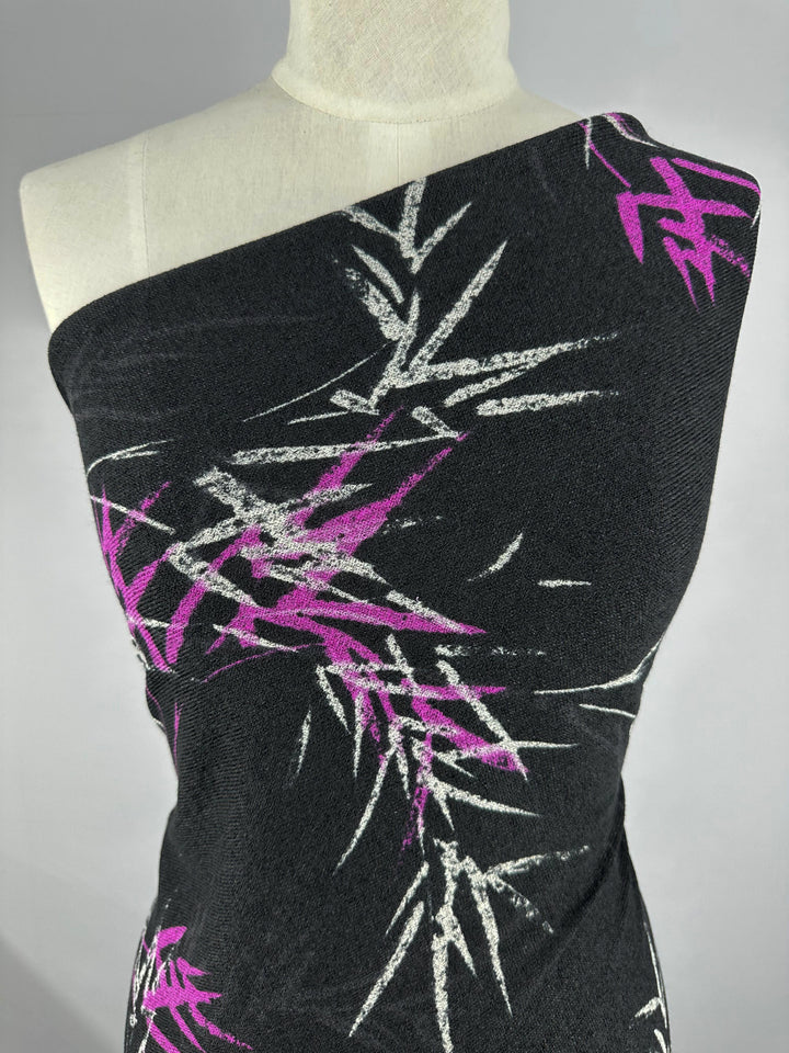 A mannequin dons the Super Cheap Fabrics Textured Knit - Bamboo dress with a black, one-shoulder design featuring a vibrant pattern of purple and white abstract bamboo leaves.