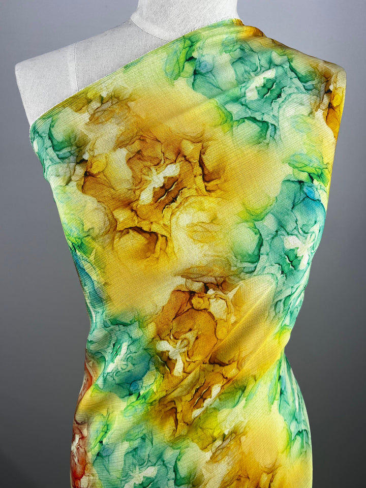 A mannequin draped with Super Cheap Fabrics' Designer Cotton - Mantis - 150cm displays a luxurious, watercolor-inspired artistic pattern featuring abstract swirls of yellow, green, and blue, accented with hints of orange and brown, perfect for versatile luxury sewing projects.