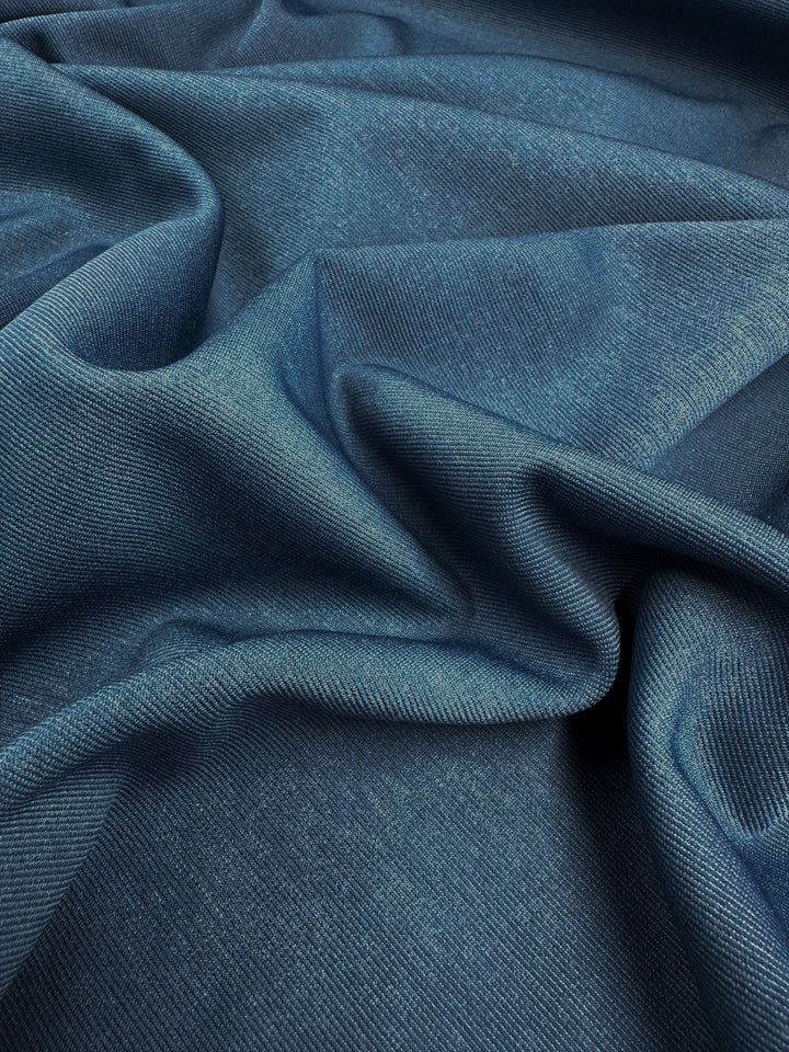 A close-up image of a piece of Ponte - Bluesteel - 157cm fabric from Super Cheap Fabrics shows the texture and folds of the material. The light creates subtle highlights and shadows, accentuating the soft, smooth surface of the wrinkle-resistant fabric.