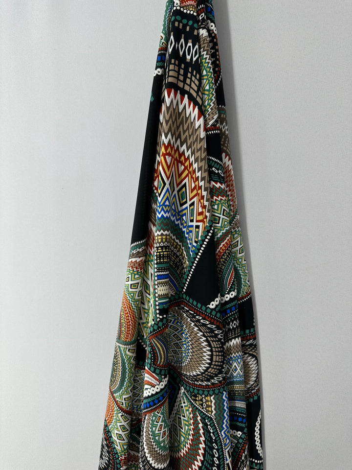 A colorful, patterned Textured Lycra - Africa - 150cm by Super Cheap Fabrics with intricate geometric designs in various colors including green, red, white, and black hangs against a plain, light gray wall. This polyester blend features a mix of zigzags, diamonds, and circular patterns.