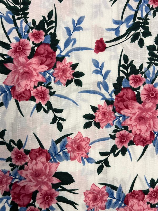 The Textured Deluxe Print - Catch The Bouquet from Super Cheap Fabrics features an intricate floral pattern with pink and red flowers, green leaves, and blue accents on a textured white base. This versatile polyester fabric is perfect for adding a harmonious and vibrant touch to designer creations.