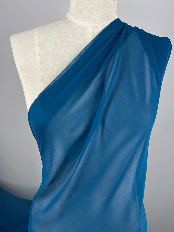 The white mannequin is elegantly draped with the Super Cheap Fabrics' Silk Georgette - Celestial - 135cm, styled asymmetrically over one shoulder. This lightweight, sheer blue fabric is ideal for spring or summer, offering a delicate glimpse of the mannequin beneath. The plain, light-colored background further accentuates the graceful drape.