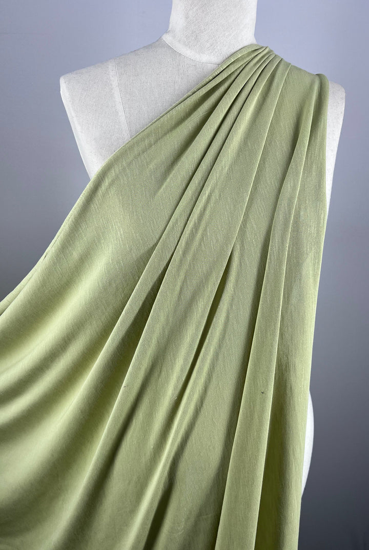 A close-up of a mannequin draped in Super Cheap Fabrics' Summer Cotton Knit - Beechnut, a lightweight, soft fabric in light green. The material is smoothly gathered over one shoulder, highlighting its texture and flow against a neutral gray background.