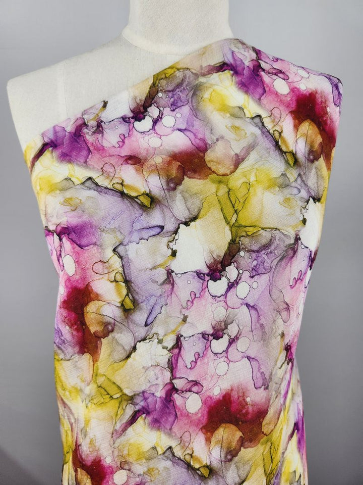 A close-up image showcases a mannequin adorned with "Designer Cotton - Blist - 150cm" from Super Cheap Fabrics. The luxurious material displays an abstract pattern of swirling purple, pink, yellow, and white hues reminiscent of watercolor art, set against a plain gray background—ideal for luxury sewing projects.