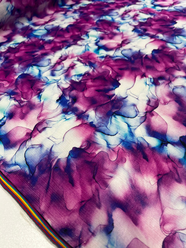 This image displays a close-up of the "Designer Cotton - Celeste - 150cm" fabric by Super Cheap Fabrics, showcasing a vibrant abstract floral design in hues of purple, blue, and white. The breathable material is soft with gathered elements that mimic layered petals. A subtle striped edge is noticeable at the bottom.