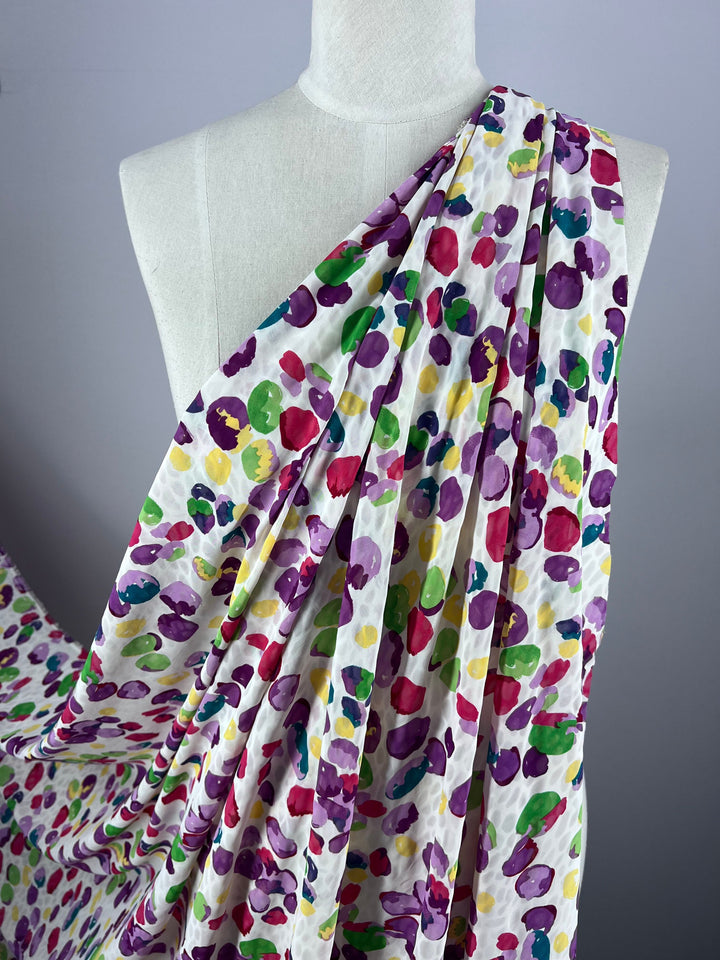 A mannequin draped in Super Cheap Fabrics' Salt Shrunk Print - Pebbly, 155cm, features a vibrant pattern of colorful dots in purple, pink, red, yellow, and green on white. This standout piece creates flowing layered folds.