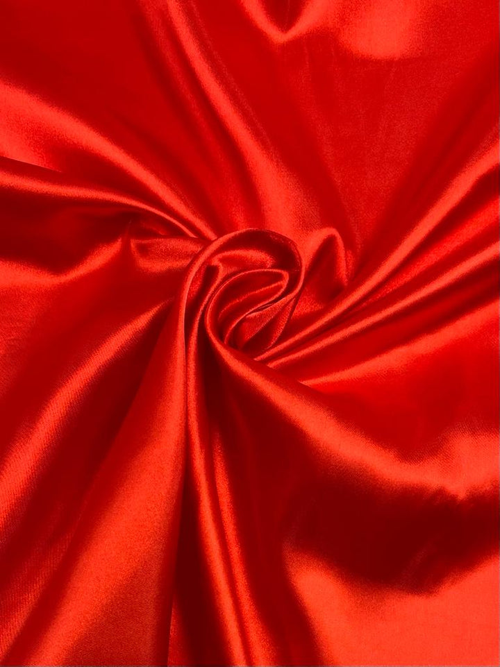 A close-up of Super Cheap Fabrics' Satin in Red (150cm) reveals its smooth, shiny texture with gentle folds spiraling toward the center. Light reflects off this vibrant fabric, enhancing its rich, glossy look—ideal for elegant dresses or luxurious home décor accents.