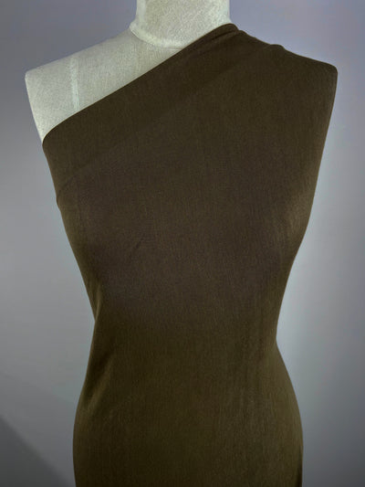 Displayed against a gray backdrop, the mannequin showcases the "Summer Cotton Knit - Sea Turtle - 140cm" by Super Cheap Fabrics—a brown, one-shoulder dress in lightweight cotton. Its smooth, fitted material highlights the simplicity and elegance of this stylish design.