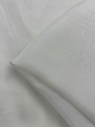 A close-up image of a pillow in platinum, made with Pure Silk Chiffon from Super Cheap Fabrics, resting on matching light gray bed sheets. The fabric appears smooth and soft, with subtle creases indicating the pillow's shape and the texture of the 100% silk material.