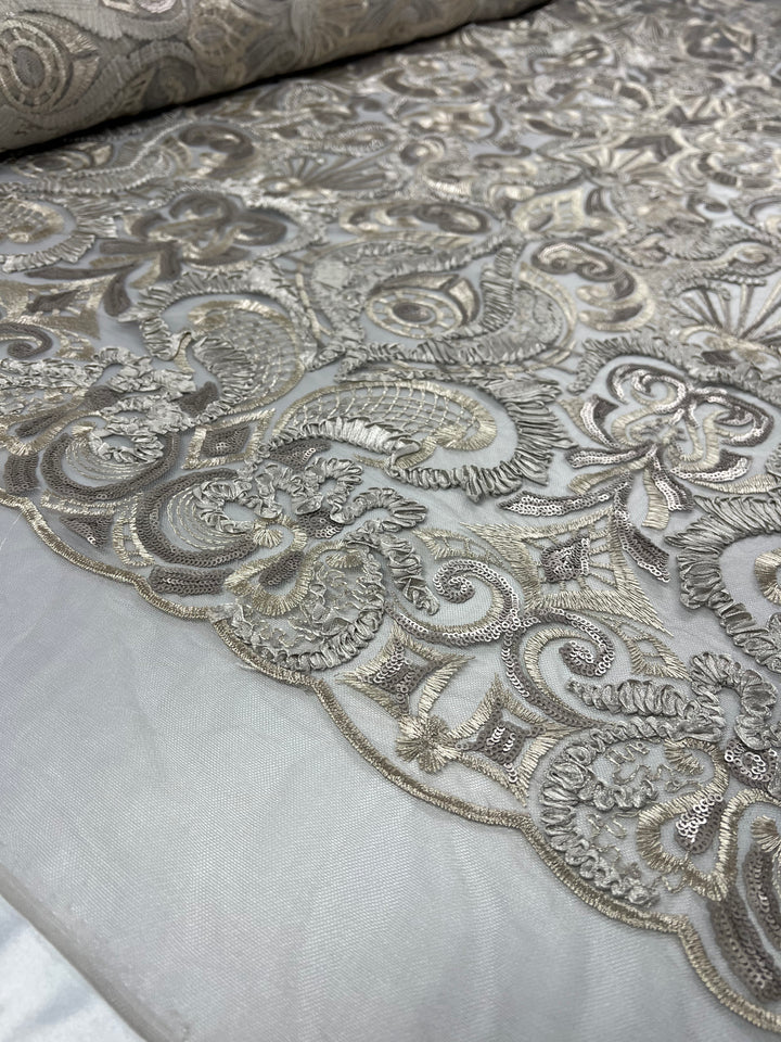 A close-up of a luxurious medium weight fabric with intricate gold and silver embroidery. The black multi fabric appears to be lace with detailed floral and scroll patterns, laid out flat and partially rolled on one end. The texture and shine of the embroidery are clearly visible. This is the Embroidery on Tulle - 150cm - Jewel - 150cm by Super Cheap Fabrics.