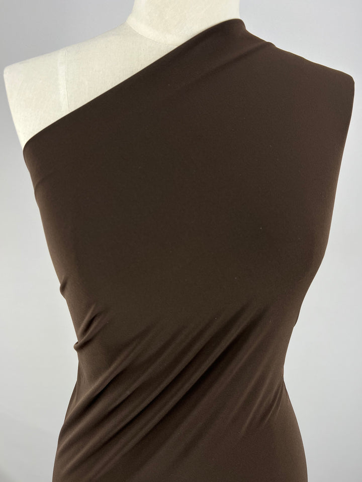 A close-up of a dress form showcasing an asymmetrical, one-shoulder brown dress made from smooth, stretchy ITY Knit - Brown - 150cm by Super Cheap Fabrics. The medium weight fabric drapes elegantly with subtle creases and a sleek finish. The background is plain and light-colored.