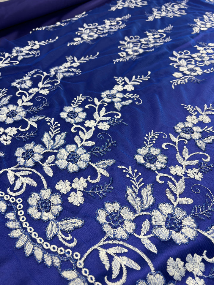 A close-up of intricate white floral embroidery on a rich, royal blue fabric. The design features various flowers, leaves, and delicate swirls, creating an elegant and ornate pattern. The lightweight fabric has a lustrous sheen, highlighting the detailed needlework. This is the Embroidered Lace - Royal - 150cm by Super Cheap Fabrics.