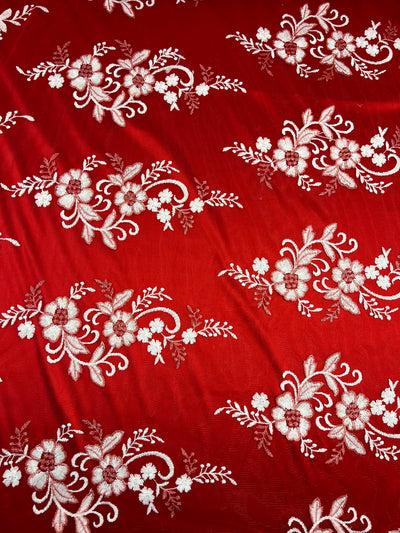 A vibrant scarlet fabric adorned with intricate white floral embroidery patterns. The design features clusters of flowers and leaves, symmetrically arranged and repeated across the polyester textile, creating an elegant and decorative appearance. This is the Embroidered Lace - Scarlet - 150cm by Super Cheap Fabrics.