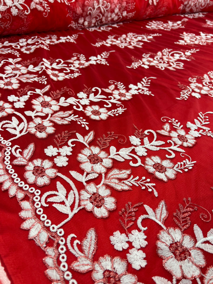 A close-up photo of vibrant scarlet fabric features intricate white floral embroidery. The detailed patterns include various flowers and leaves, creating a delicate and elegant design on the richly colored polyester material, which is known as the Embroidered Lace - Scarlet - 150cm from Super Cheap Fabrics.