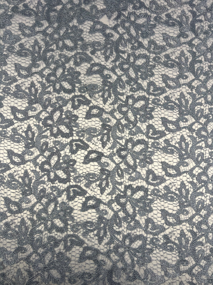 A close-up of Super Cheap Fabrics' Evening Lace - Metallic Blue - 120cm sheer polyester fabric with an intricate lace pattern. The lace features an ornate floral design with leaves and vines woven together, creating an elegant and delicate appearance. The background appears to be a light, neutral color, enhancing the lace details.