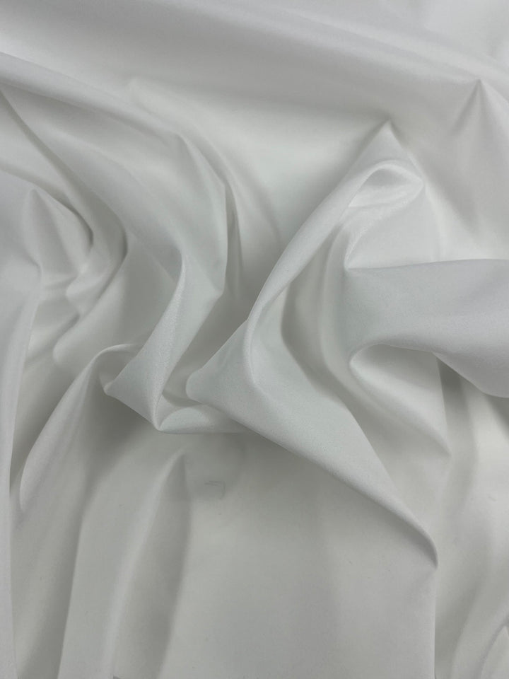 A close-up image of smooth, white fabric lining with soft folds and creases. The material appears light and slightly glossy, creating a subtle play of shadows and highlights on its surface, showcasing the expert tailoring in its design. This is the Lining - White - 150cm from Super Cheap Fabrics.