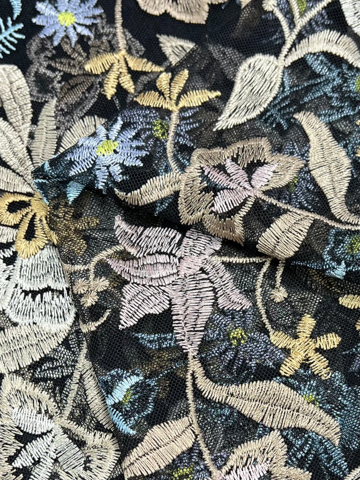 Close-up of intricately embroidered mesh fabric, featuring a variety of floral and leaf patterns. The Embroidered Lace - Dolce - 150cm by Super Cheap Fabrics, crafted with polyester threads in pastel shades of pink, yellow, and blue on a dark background, creates a textured and detailed design. The fabric appears slightly folded.