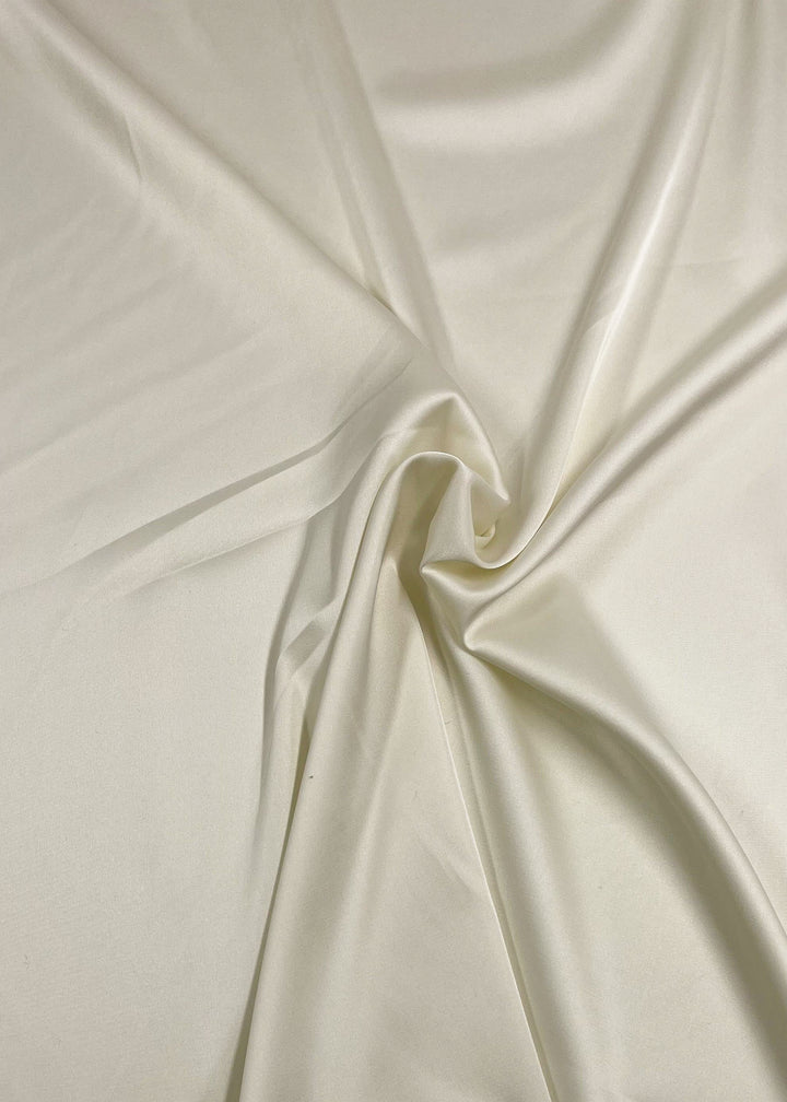 A close-up image of a piece of Satin Deluxe - Ivory - 150cm by Super Cheap Fabrics that appears to have a soft, shiny finish. The material is slightly wrinkled and gathered in the center, creating gentle folds and smooth textures throughout. The satin reflects light beautifully, giving it a glossy appearance.