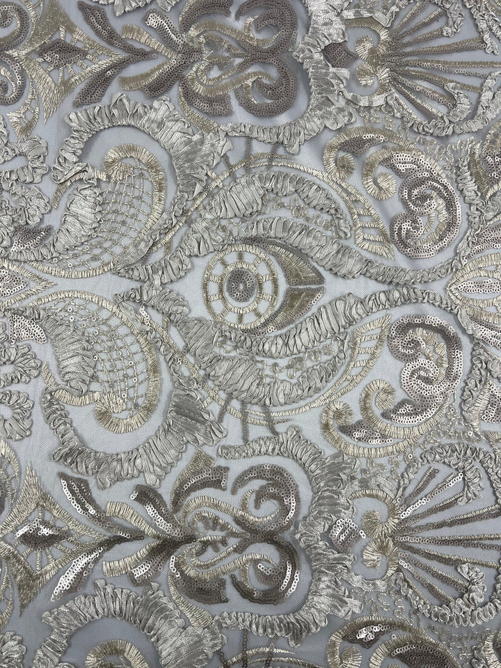 A close-up image of intricate silver and gray embroidery on medium weight fabric. The pattern includes swirling lines, floral shapes, and textured designs, creating an elaborate and ornate appearance. The delicate stitching gives the fabric a luxurious and detailed look. This is Embroidery on Tulle - 150cm - Jewel - 150cm from Super Cheap Fabrics.