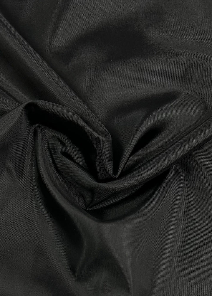 A close-up image displaying a piece of Lining - Black - 150cm by Super Cheap Fabrics with a glossy finish. The lightweight lining fabric appears to be draped and folded, creating smooth, flowing lines and subtle shadows, showcasing its texture and sheen.