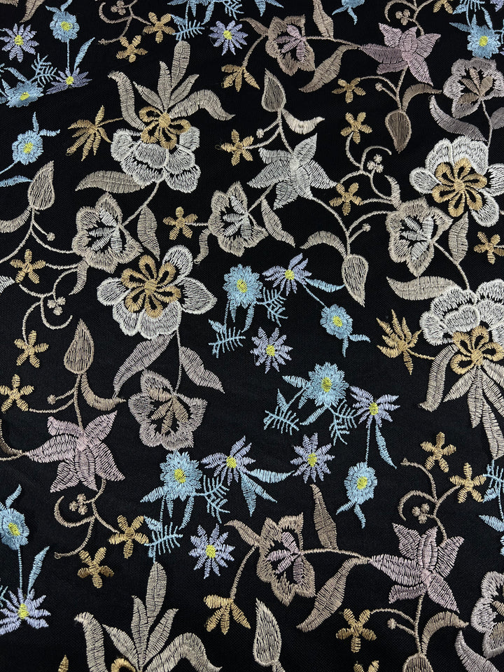 A detailed embroidery design on mesh fabric featuring an array of flowers and foliage. The flowers are in pastel shades of white, pink, blue, and yellow, with intricate and intertwined stems and leaves, showcasing a delicate and ornate pattern. This is the Embroidered Lace - Dolce - 150cm from Super Cheap Fabrics.