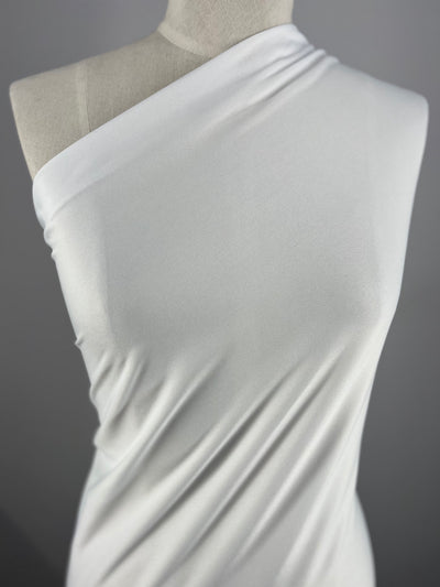 A close-up of a mannequin draped in a smooth, white, 100% polyester one-shoulder fabric. The lightweight Interlock - White - 150cm from Super Cheap Fabrics appears to be soft and slightly reflective, creating gentle folds that emphasize its luxurious texture. The background is a neutral gray.