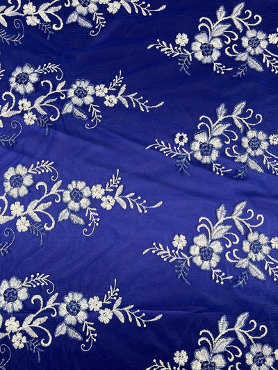 A section of Super Cheap Fabrics' Embroidered Lace - Royal - 150cm adorned with intricate white floral embroidery. The embroidered flowers, leaves, and stems are arranged in a repeating pattern, creating an elegant and detailed design.