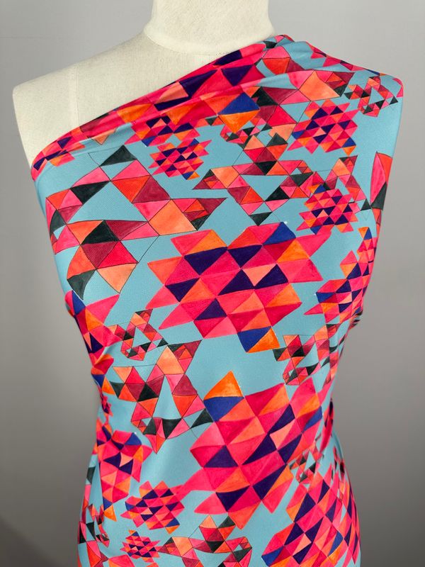 A mannequin is draped in a brightly colored Printed Lycra - Geo Village - 150cm fabric from Super Cheap Fabrics. This medium-weight material features vibrant, overlapping triangles and diamond shapes in shades of pink, red, blue, and purple on a light blue background. The garment is one-shouldered.