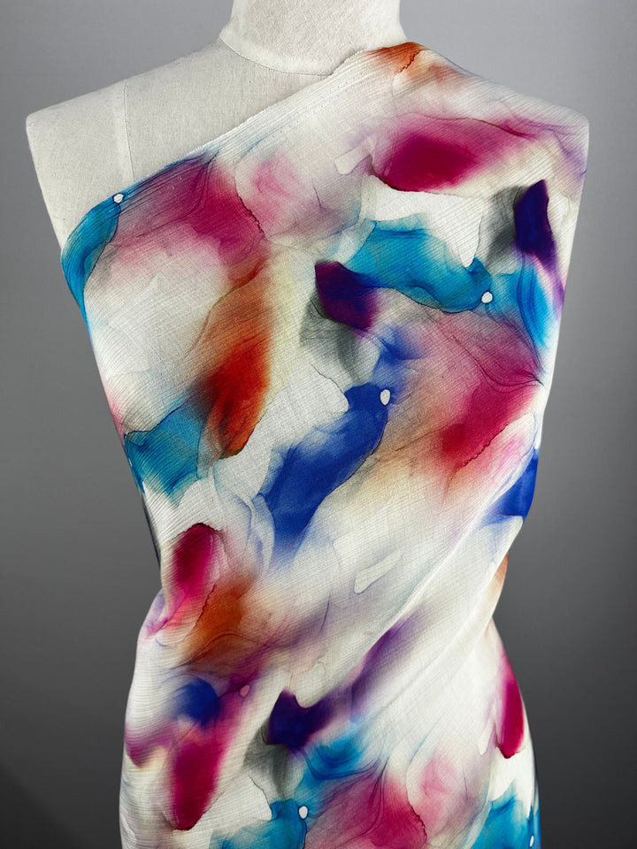 A mannequin draped with Super Cheap Fabrics' Designer Cotton - Crysta - 150cm showcases an abstract watercolor pattern. The vibrant shades of blue, pink, orange, and white blend seamlessly across the cloth. Ideal for luxury sewing projects, it stands against a plain gray background.