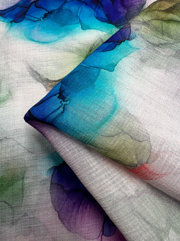 This close-up showcases the Designer Cotton - Azurine - 150cm from Super Cheap Fabrics, adorned with an abstract watercolor design in vibrant shades of blue, green, and purple, complemented by subtle red accents. Perfect for luxury sewing projects, the fabric's soft texture and gentle folds provide a beautifully flowing appearance.