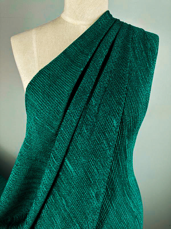 A mannequin draped in an elegant, deep teal textured fabric from Super Cheap Fabrics showcases the luxurious Metallic Knit - Amazon - 150cm. The medium to heavy weight knit is arranged in a pleated, one-shoulder design, highlighting its intricate patterns and opulent material. Set against a plain grey background, the details of this stunning fabric are perfectly emphasized, making it ideal for creating eye-catching tops or dresses.