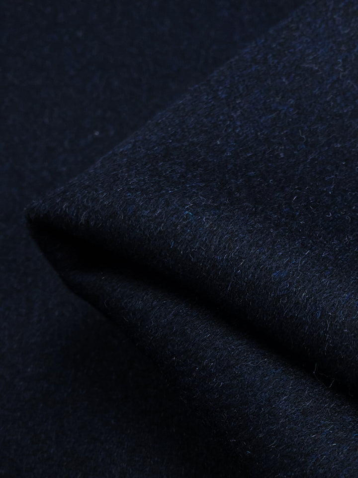 A close-up image of the Wool Cashmere - Night Sky fabric from Super Cheap Fabrics showcases its rich dark blue color, reminiscent of a night sky, with a soft and fuzzy texture punctuated by slightly visible fibers. The 150cm-wide fabric is folded, creating a subtle, angled crease that highlights variations in shading from the lighting. This elegant fabric is dry clean only.