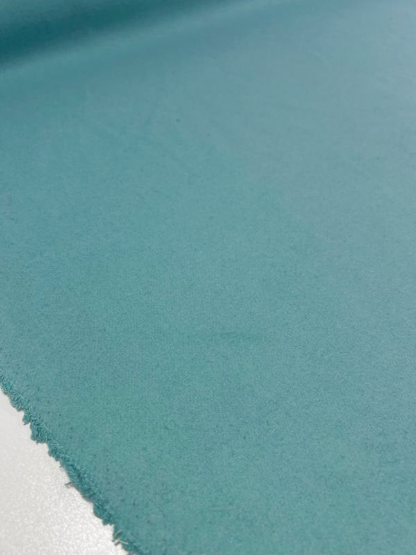 A close-up view of the Wool Cashmere - Aqua Sea fabric from Super Cheap Fabrics, displaying its light teal hue and extra-heavy weight. The material, with a slightly fuzzy texture, is lying flat on a surface and features a smooth appearance with a mildly frayed edge on the bottom left, lending it a casual, unfinished look. This fabric is ideal for outer coats and should be dry cleaned only.