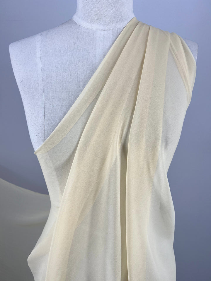 A close-up image of a mannequin draped in soft, sheer beige fabric. The Silk Georgette - Chardonay from Super Cheap Fabrics is arranged to cover one shoulder, falling gracefully down the front and back, creating elegant folds and an airy appearance. Perfect for summer outfits.