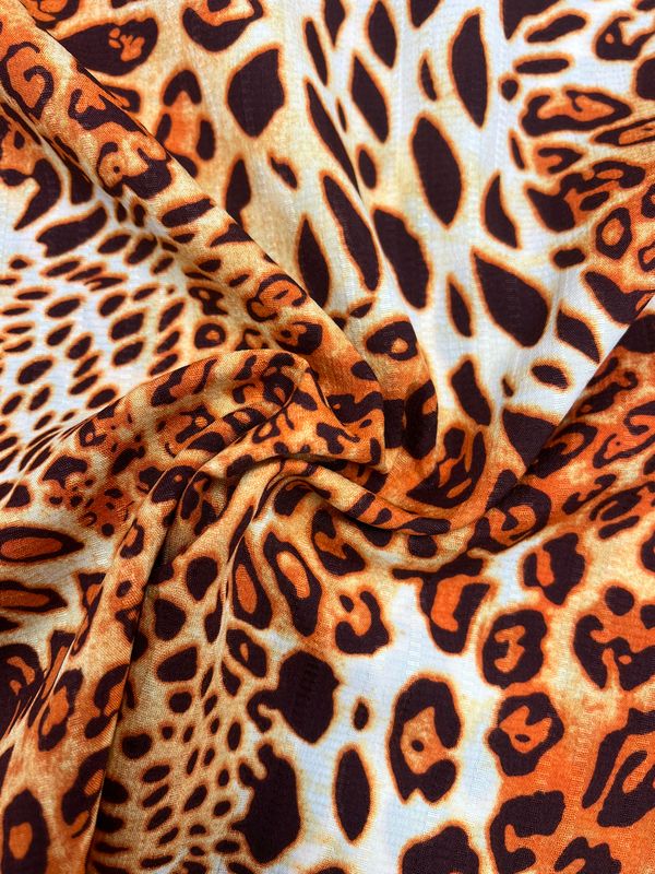 A close-up view of the Textured Deluxe Print - Leo - 155cm by Super Cheap Fabrics reveals vibrant fabric adorned with a bold leopard print pattern. This design boasts orange and black spots of varying sizes on a light tan background, creating dynamic and engaging textures and colors. The slightly wrinkled polyester material adds depth to this designer fabric, enhancing its visual appeal.