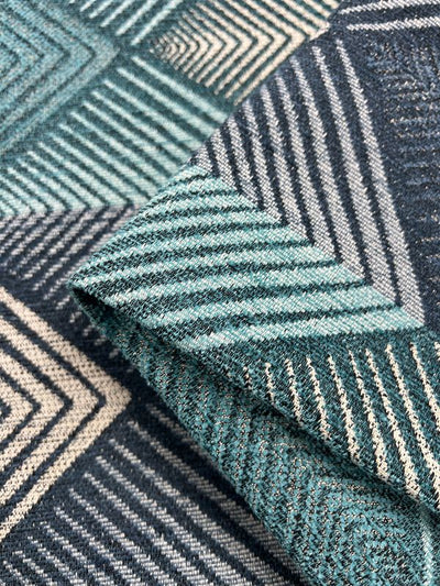 Close-up of Upholstery Jacquard - Ocean Ray - 143cm by Super Cheap Fabrics, featuring teal, white, and dark blue triangular and diamond shapes. The durable fabric is slightly folded, showcasing the intricate design and texture, perfect for enhancing your furniture.