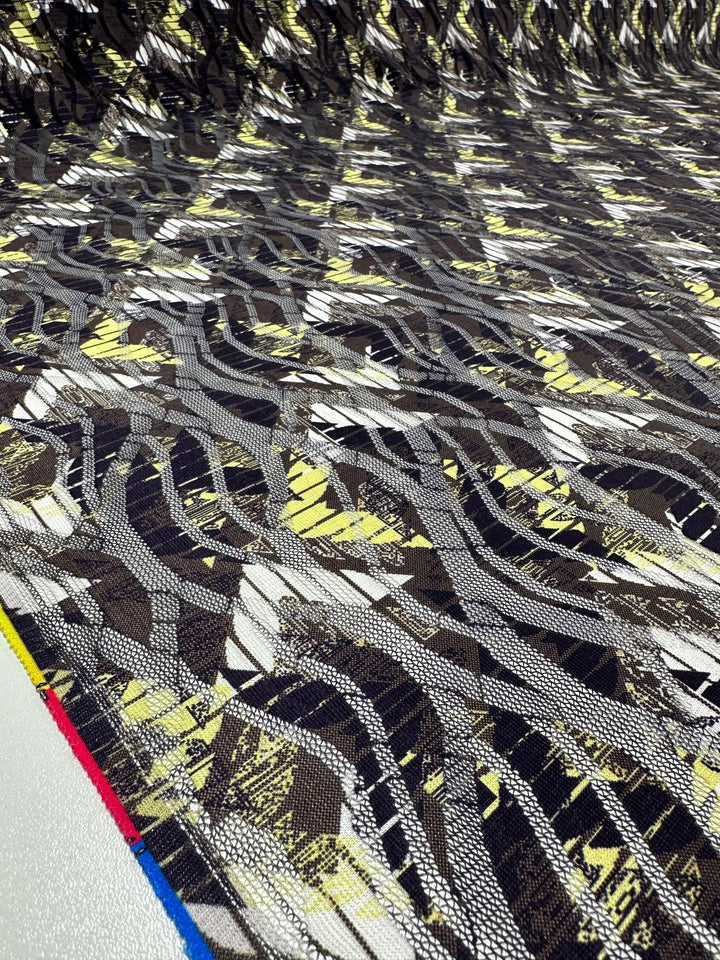 The Designer Mesh - Direction - 150cm by Super Cheap Fabrics is a textured abstract polyester with overlapping wavy patterns in gray, black, white, and yellow. It has a layered look and features small red, blue, and yellow bordered sections on one edge—ideal for home décor accents.