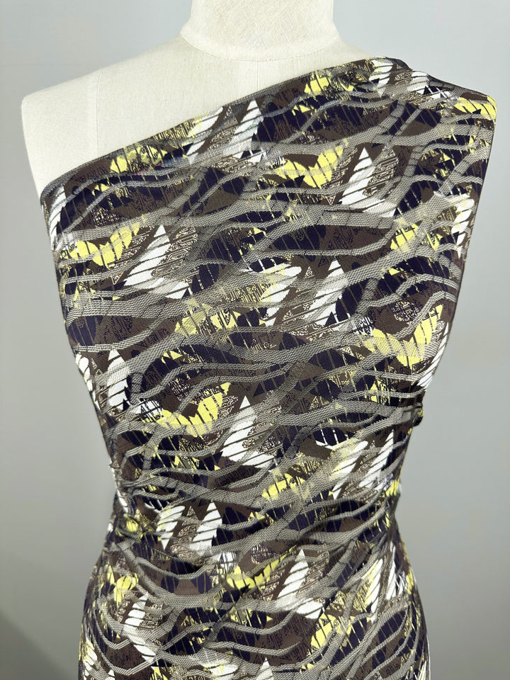 A mannequin displays a Designer Mesh - Direction dress by Super Cheap Fabrics, featuring an abstract leaf pattern in brown, yellow, white, and black. Crafted from smooth polyester fabric, it drapes elegantly for a sleek and modern look.