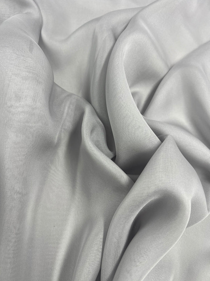 Close-up image of Super Cheap Fabrics' Pure Silk Chiffon in Silver, measuring 140cm wide. The 100% silk fabric showcases a smooth and slightly shiny texture, gently draped and folded to create soft waves and subtle shadows. The material appears soft and silky.