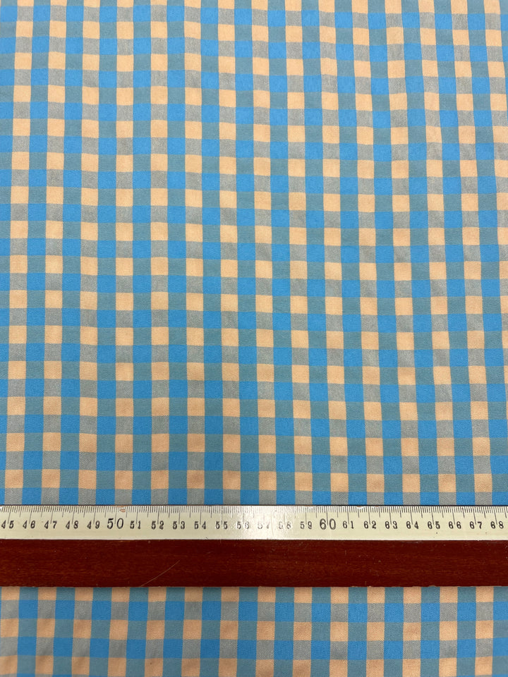 A close-up of the light weight "Cotton Poly - Cream & Blue Gingham - 150cm" fabric by Super Cheap Fabrics, showcasing its blue and caramel cream checkered pattern. A ruler with centimeter markings is placed horizontally across the bottom part of the fabric, making it ideal for crafting stylish pants and skirts.