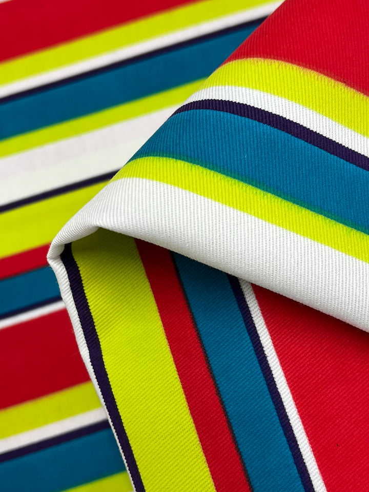 A close-up photo of the Printed Denim - Temperature Stripes from Super Cheap Fabrics. The pattern features horizontal stripes in vibrant hues of red, blue, yellow, green, and black on a white background. The 100% cotton fabric is partially folded, highlighting its bold design. This light medium weight fabric radiates multi-color brilliance across its 150cm width.