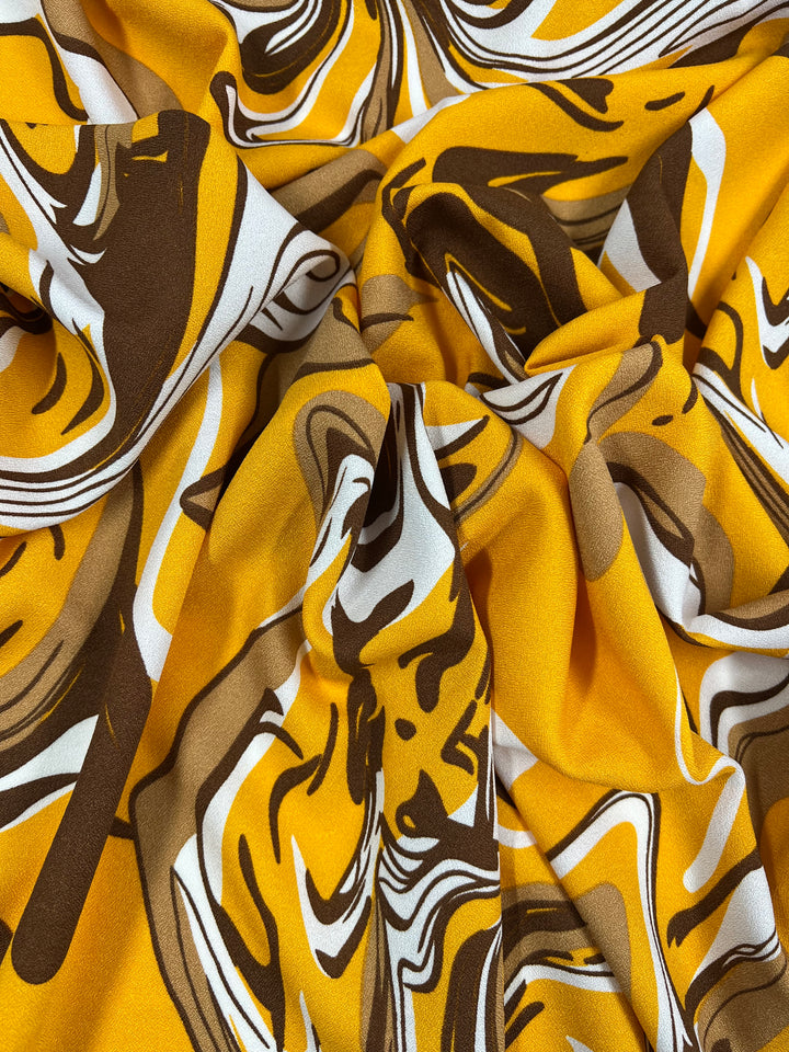 A close-up shot of a crumpled piece of Printed Crepe fabric from Super Cheap Fabrics showcases a vibrant abstract pattern in yellow, brown, black, and white. This multi-colored design on the Cappuccino variant with a width of 150cm creates a dynamic, artistic appearance.